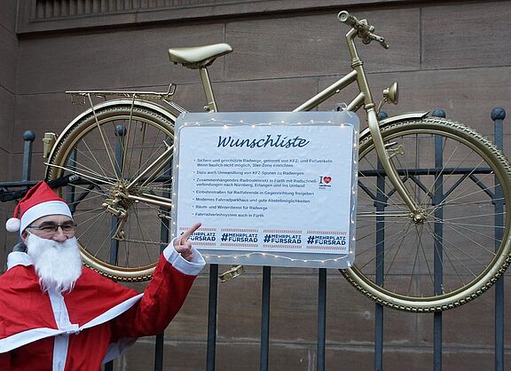 Ein Mensch im Weihnachtsmannkostüm zeigt auf eine Liste, die an einem Fahrrad befestigt ist. Es hängt am Zaun und enthält Wünsche für sichere und geschützte Radwege.
