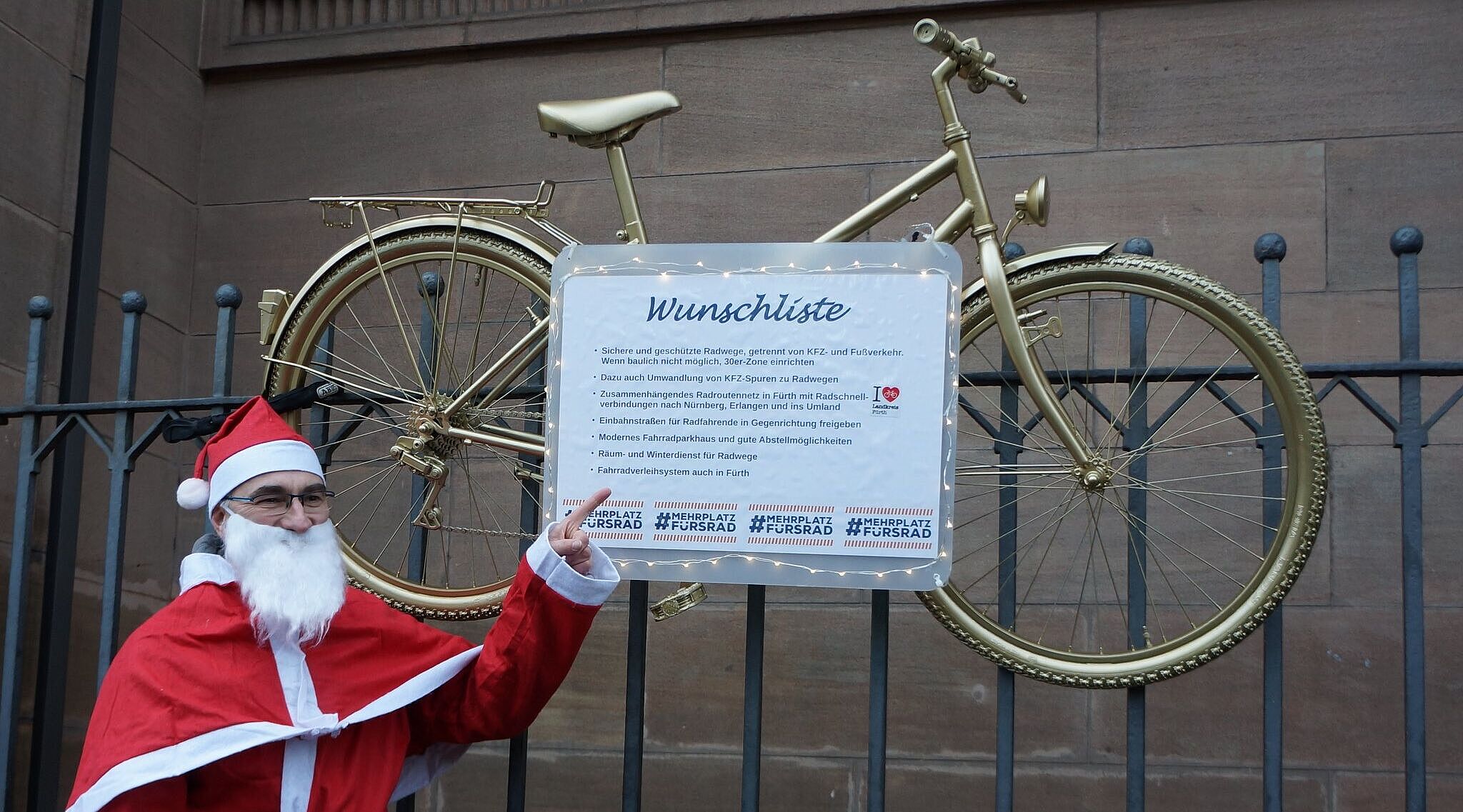 Ein Mensch im Weihnachtsmannkostüm zeigt auf eine Liste, die an einem Fahrrad befestigt ist. Es hängt am Zaun und enthält Wünsche für sichere und geschützte Radwege.