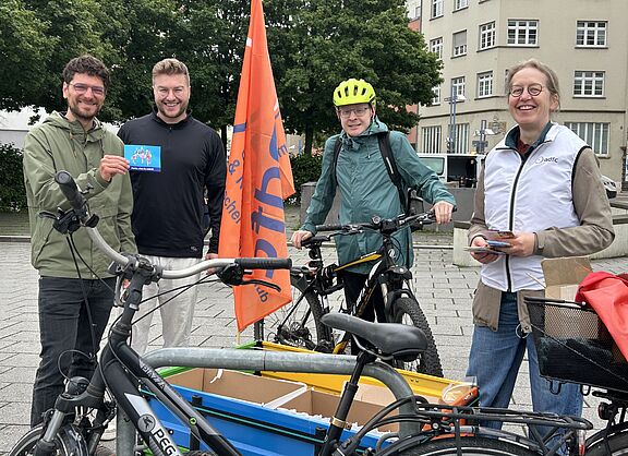 Drei Männer und eine Frau mit Fahrrädern im Vordergrund danken den Radfahrenden zum Weltfahrradtag.mit einem kleinen Geschenk zum 