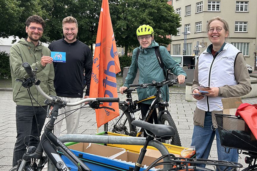 Fahrrad-Dankstelle Drei Männer und eine Frau mit Fahrrädern im Vordergrund danken den Radfahrenden zum Weltfahrradtag.mit einem kleinen Geschenk zum
