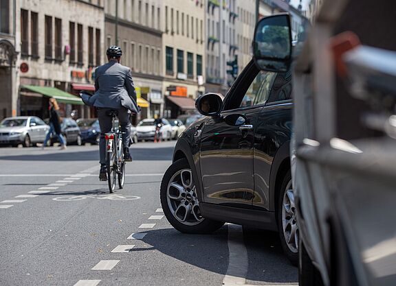 Radverkehr im Stadtverkehr Radfahrer in dunkler Kleidung mit Helm fährt auf einer Stadtstraße, im Vordergrund steht ein schwarzes Auto am Straßenrand, weitere Fahrzeuge und Gebäude im Hintergrund.