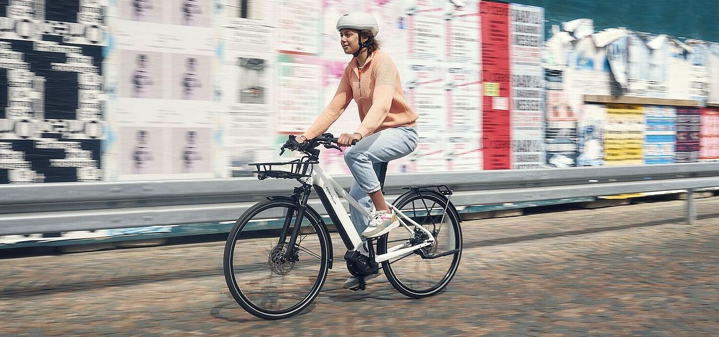 Eine Frau fähet auf einem weißen Elektrorad vor einer Wand mit vielen Plakaten. 