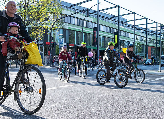 Eine Gruppe von Kindern fährt auf ihrer Rädern auf der Straße. Sie werden von Erwachsenen begleitet.